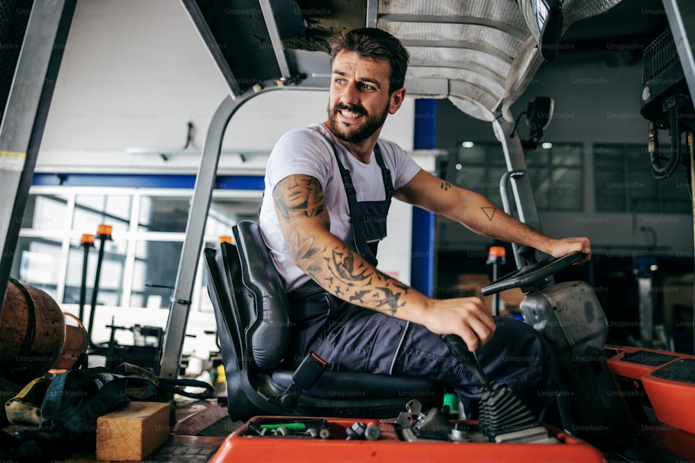 Smiling bearded tattooed hardworking employee in overalls sitting in vehicle and driving it.