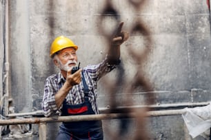 A senior factory worker in overalls, with helmet, speaking on talkie-walkie with coworkers and pointing where to put freight.