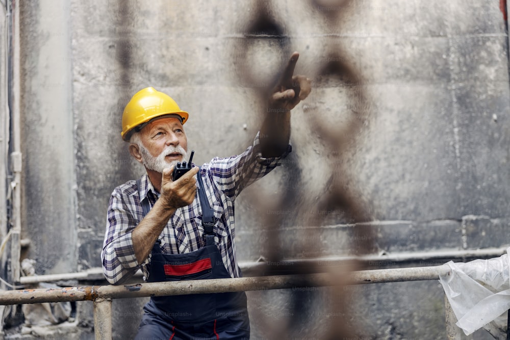 A senior factory worker in overalls, with helmet, speaking on talkie-walkie with coworkers and pointing where to put freight.
