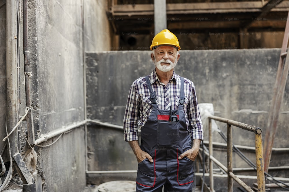 An old but experienced factory worker with a helmet on his head is standing in the factory and holding hands in his pockets. He is ready for new tasks.