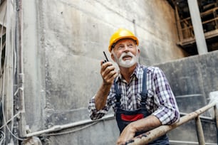 An old factory worker with a helmet on his head, leans on the railing inside of the factory and communicates with his coworkers over the walkie-talkie. Workers talking with walkie-talkie.