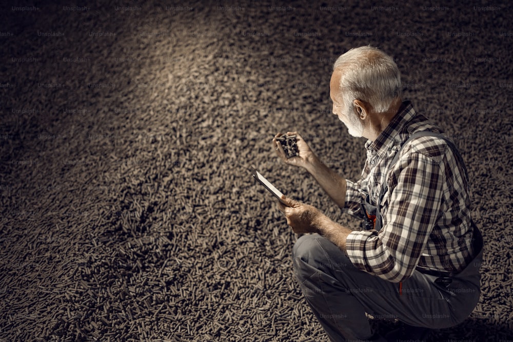 Experience, technology, and knowledge in the sugar industry, A senior supervisor is crouching in the dark room with sugar beet products, holding some of them and checking on a tablet if it is good.