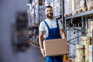 A happy storage worker in overalls with a tattoo on his arm is relocating a box with goods while walking in storage. The box is ready for shipping.