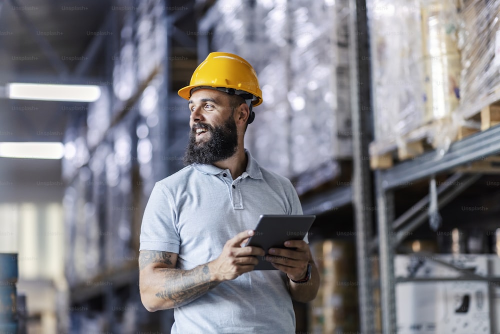 An inspector with a helmet on his head, stands in the warehouse and checks on inventory on the tablet. There are all around goods on shelves.