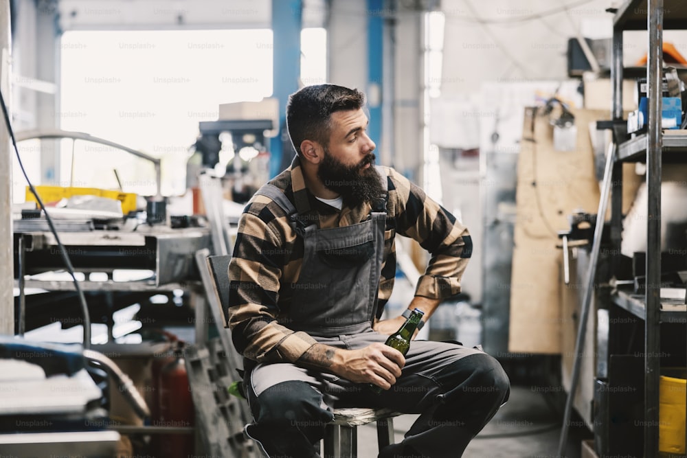 A factory handyman sits in a facility with a bottle of beer and taking a break.
