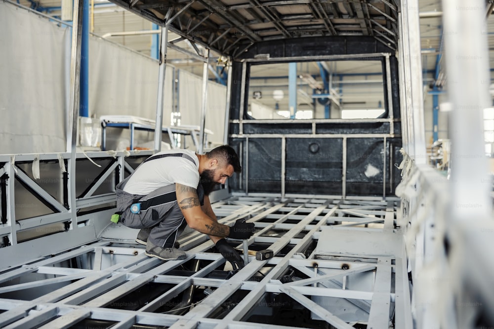 A metal worker crouching in skeleton of a new bus and building it at workshop.