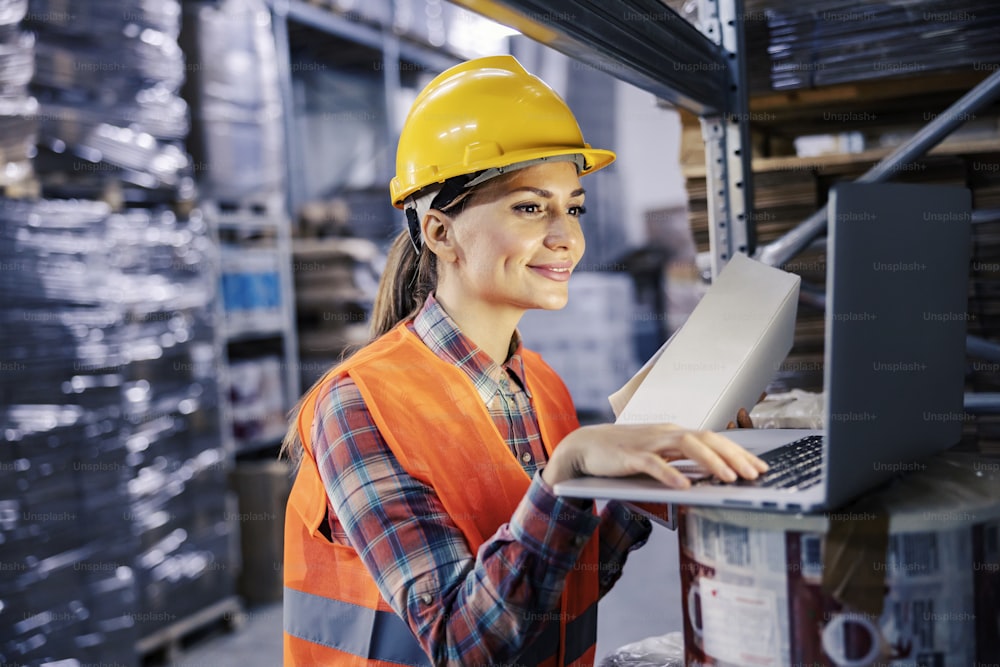 A female storage worker adding order on list while smiling at laptop.