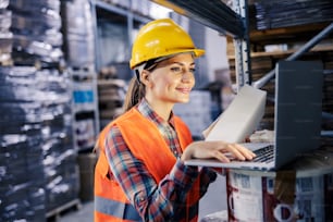 A female storage worker adding order on list while smiling at laptop.