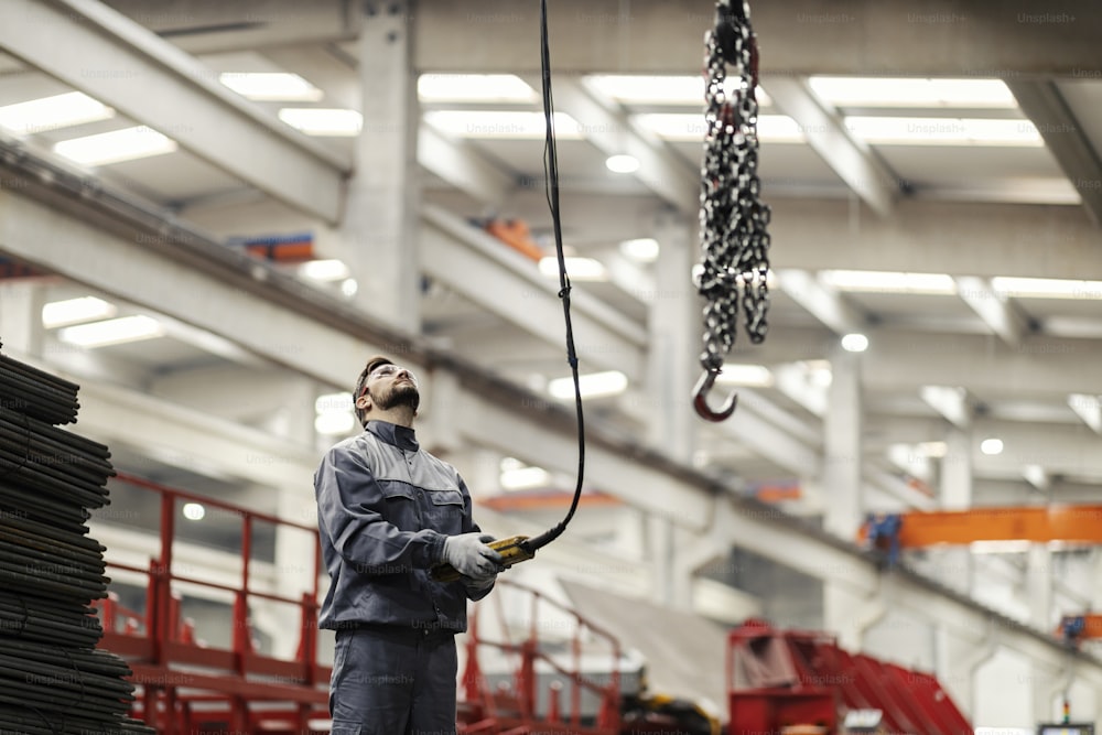 A heavy industry worker relocates chains on an industrial hook by using a controller while standing in the factory.