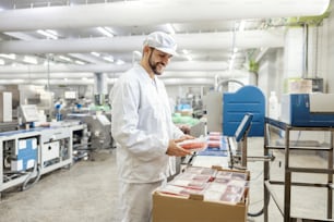 Un feliz trabajador de una fábrica de carne está empacando carne picada en una caja y preparándola para su entrega mientras está de pie junto a una cinta transportadora.