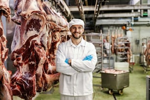 A butcher is standing by the big pieces of meat in slaughter house and smiling at the camera.