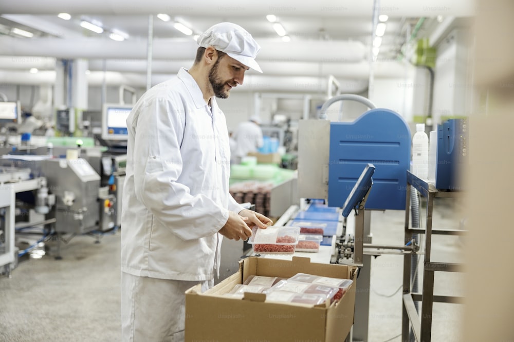 A meat industry laborer is packing minced meat into a box and getting ready to deliver it while standing next to a conveyor belt.