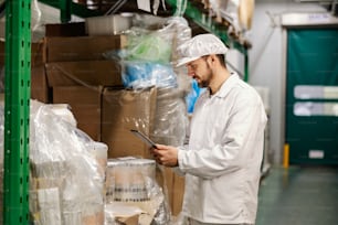 A food factory worker is inventorying on tablet while standing in factory storage.