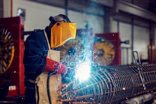 A heavy industry metal worker is welding metal framework with a welding machine in a factory.