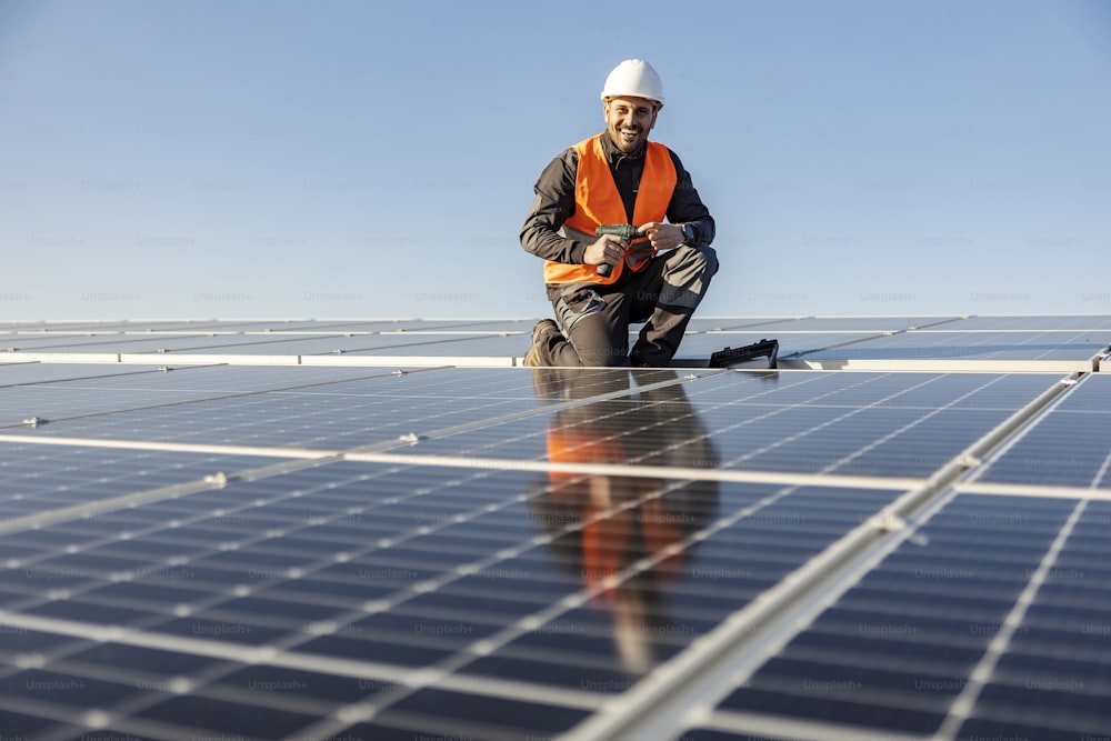 Ein Arbeiter, der in die Kamera lächelt und einen Schraubenzieher in der Hand hält, um Sonnenkollektoren auf dem Dach zu installieren.
