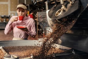 A female factory supervisor drinking aromatic cup of coffee next to a roasting machine.