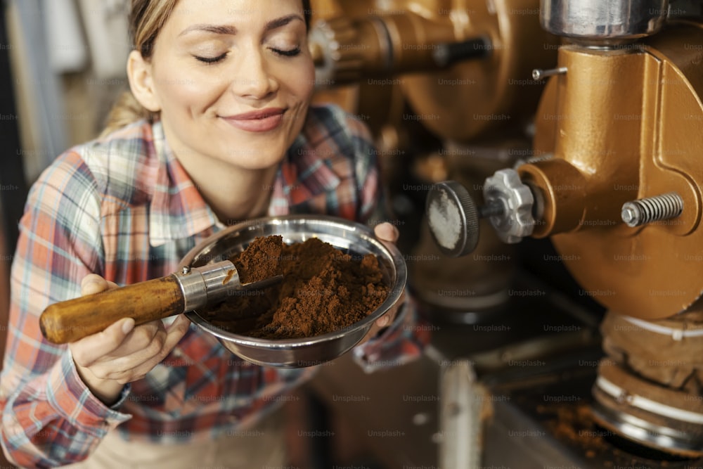 A female factory worker smelling ground fresh coffee while standing next to a machines.