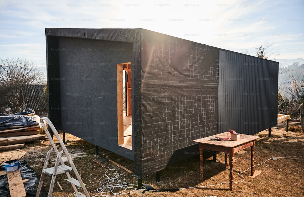 Wooden frame house on pile foundation in the Scandinavian style barnhouse under construction. Wall with waterproof membrane and black corrugated iron sheet used as facade of future cottage.