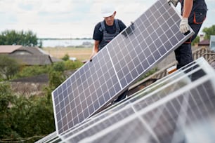 Männliche Techniker, die photovoltaische Solarmodule auf dem Dach des Hauses montieren. Bauherren in Helmen, die ein Solarpanel-System im Freien installieren. Konzept alternativer und erneuerbarer Energien.