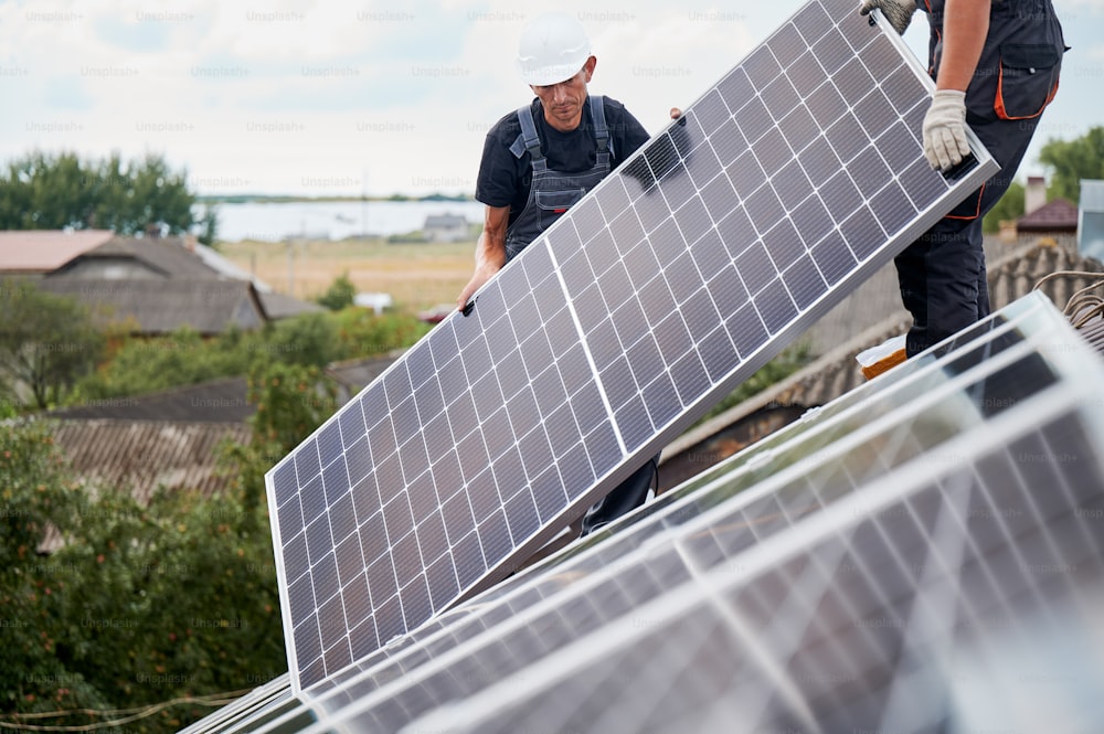 Hombres técnicos montando módulos solares fotovoltaicos en el techo de la casa. Constructores con cascos instalando sistema de paneles solares al aire libre. Concepto de energía alternativa y renovable.