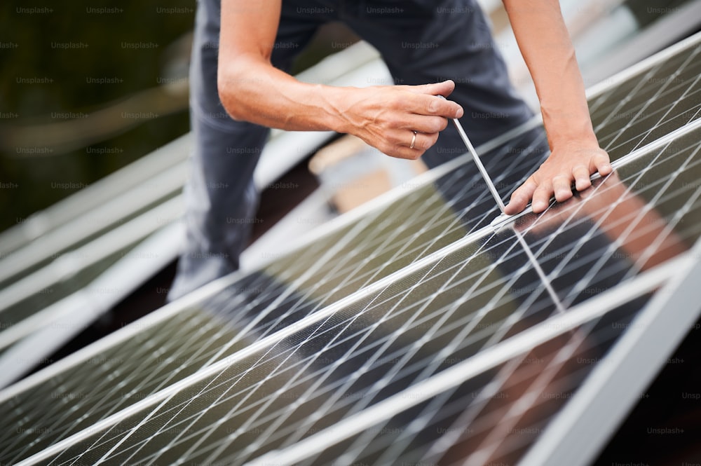 Hombre trabajador montando módulos solares fotovoltaicos en el techo de la casa. Primer plano del electricista instalando el sistema de paneles solares en exteriores, apretando con llave hexagonal. Concepto de energía alternativa y renovable.