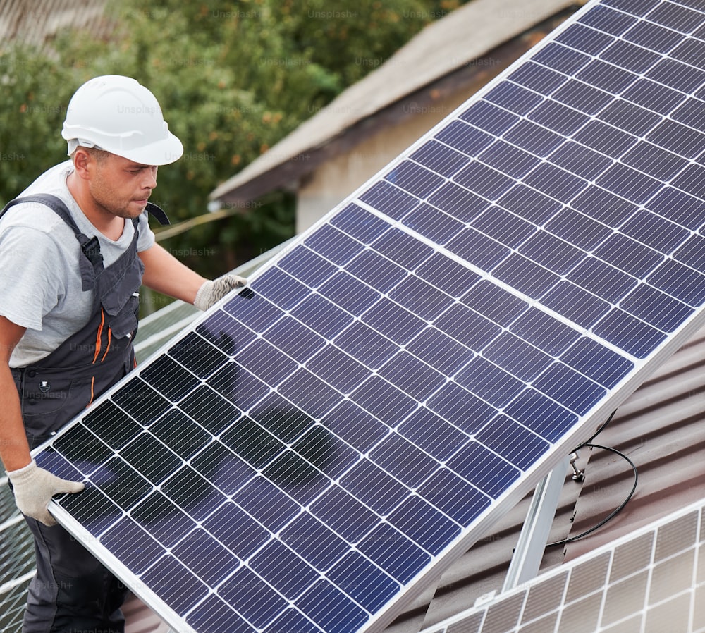 Tecnico dell'uomo che trasporta moduli solari fotovoltaici sul tetto della casa. Elettricista in casco che installa il sistema di pannelli solari all'aperto. Concetto di energia alternativa e rinnovabile.