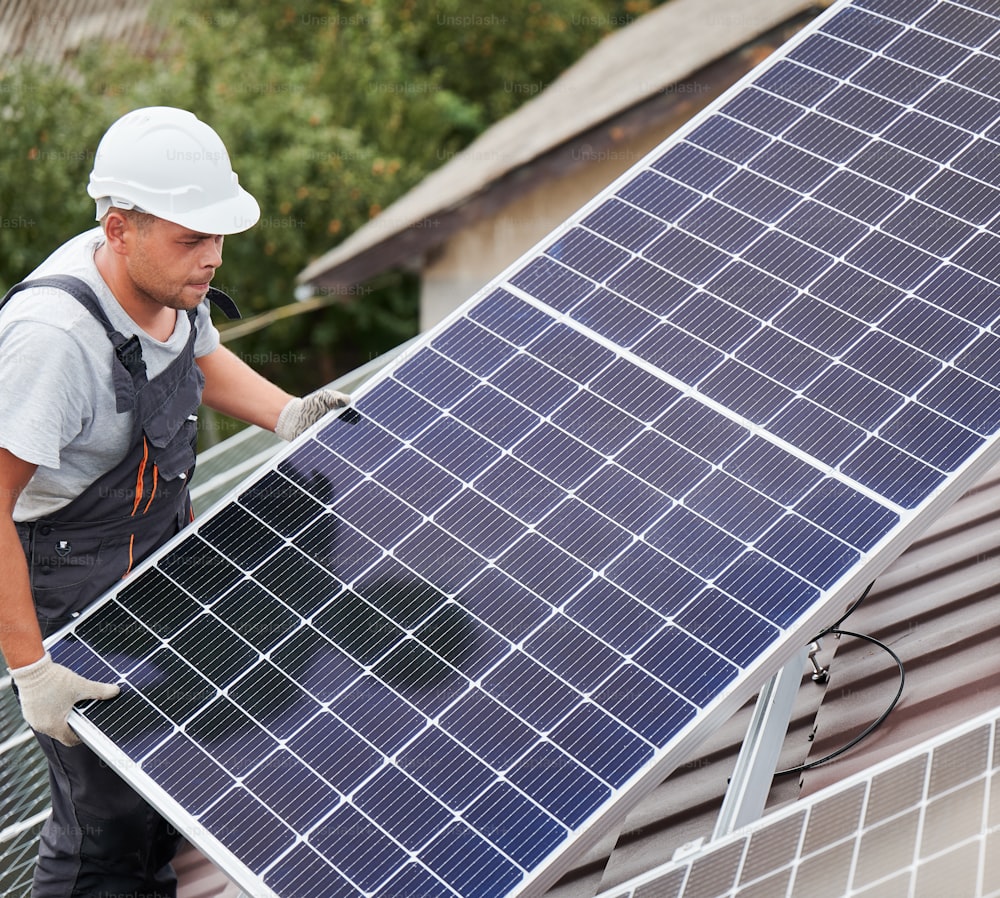 Man technician carrying photovoltaic solar moduls on roof of house. Electrician in helmet installing solar panel system outdoors. Concept of alternative and renewable energy.