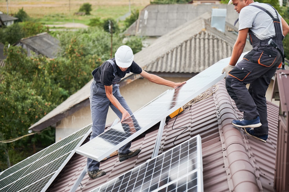 Hombres técnicos que llevan módulos solares fotovoltaicos en el techo de la casa. Constructores con cascos instalando sistema de paneles solares al aire libre. Concepto de energía alternativa y renovable.