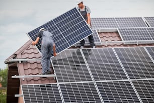 Hombres trabajadores montando módulos solares fotovoltaicos en el techo de la casa. Electricistas con cascos instalando sistema de paneles solares al aire libre. Concepto de energía alternativa y renovable.