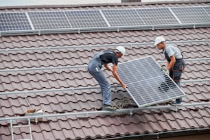 Hombres trabajadores que llevan módulos solares fotovoltaicos en el techo de la casa. Electricistas con cascos instalando sistema de paneles solares al aire libre. Concepto de energía alternativa y renovable.