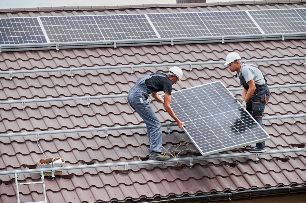Operai che trasportano moduli solari fotovoltaici sul tetto della casa. Elettricisti in caschi che installano sistemi di pannelli solari all'aperto. Concetto di energia alternativa e rinnovabile.
