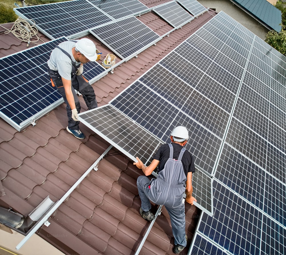 Uomini installatori che installano moduli solari fotovoltaici sul tetto della casa. Ingegneri in caschi che costruiscono sistemi di pannelli solari all'aperto. Concetto di energia alternativa e rinnovabile. Veduta aerea.