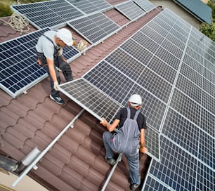 Hommes installateurs installant des modules solaires photovoltaïques sur le toit de la maison. Ingénieurs en casques construisant un système de panneaux solaires à l’extérieur. Concept d’énergie alternative et renouvelable. Vue aérienne.