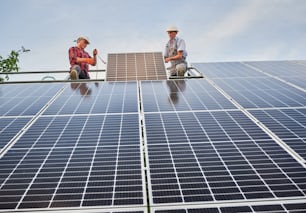 Male workers mounting photovoltaic solar panel system outdoors. Men engineers placing solar module on metal rails, wearing construction helmets and work gloves. Renewable and ecological energy.