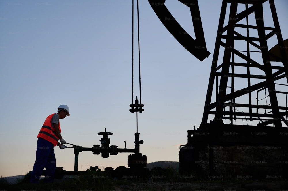 Operatore petrolifero in giubbotto da lavoro che utilizza la pompa dell'olio rocker-machine per estrarre il petrolio greggio dal pozzo. Lavoratore maschio che opera il martinetto della pompa petrolifera mentre lavora nel campo petrolifero. Concetto di estrazione dell'olio.