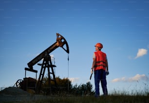 Back view of male worker in protective helmet standing near petroleum pump jack under blue sky. Petroleum engineer in work vest holding industrial wrench and looking at oil pump rocker-machine.