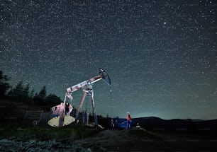 Hombre petrolero parado cerca de la bomba de petróleo Jack bajo un hermoso cielo nocturno con estrellas. Ingeniero petrolero controlando el trabajo de la máquina balancín de la bomba de aceite por la noche en el campo petrolero. Concepto de extracción de aceite.
