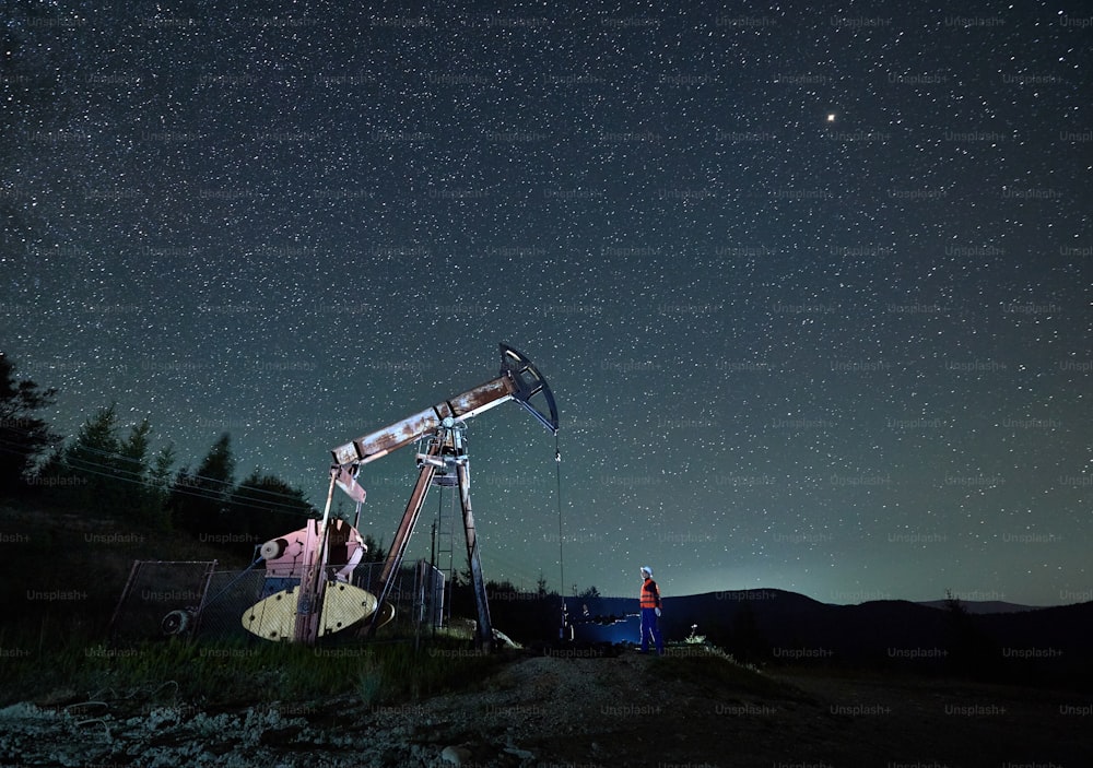 Oil man standing near petroleum pump jack under beautiful night sky with stars. Petroleum engineer controlling work of oil pump rocker-machine at night in oil field. Concept of oil extraction.