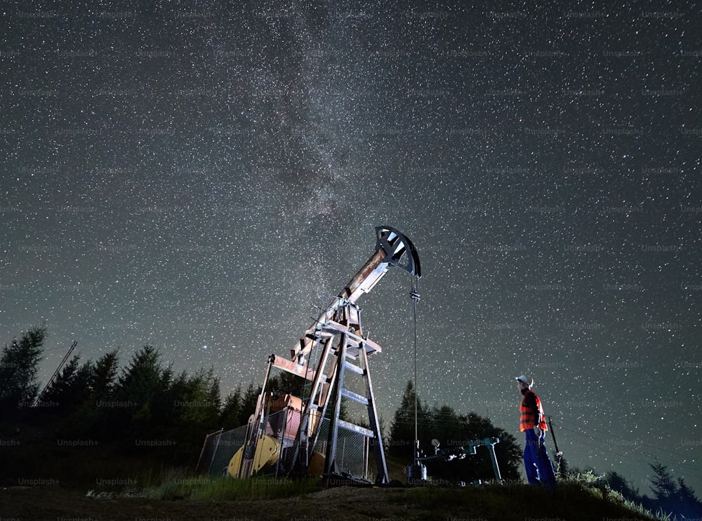 Side view of professional technician in uniform and helmet looking at derrick pump jack during night shift at oil station, male inspector checking petroleum extraction during offshore maintenance