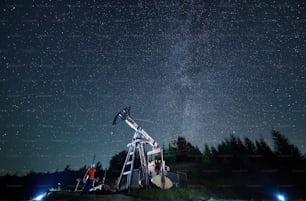 Back view of professional technician in uniform and helmet looking at derrick pump jack during night shift at oil station, male inspector checking petroleum extraction during offshore maintenance