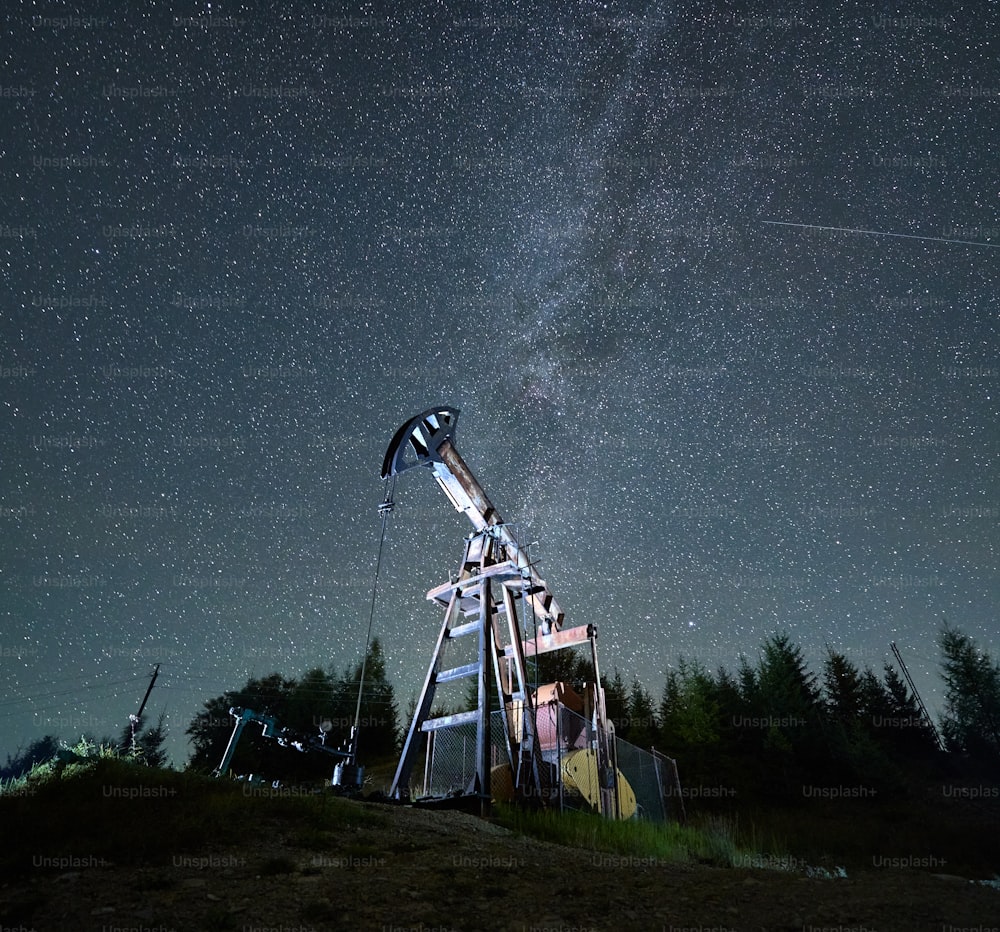 Low view of active oil well at night under beautiful starry sky. Oil and gas engineering, fuel energy. Equipment for crude oil production from fields.