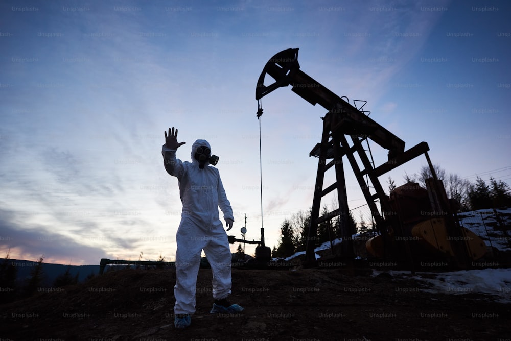 Silhouette of male scientist showing stop sign while standing on territory of oil field with pump jack and beautiful sky on background. Environmentalist wearing white radiation suit, gas mask, gloves