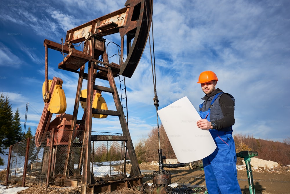 Worker holding plan of oil field and smiling to the camera at petroleum pump jack, wearing work uniform and helmet, under beautiful cloudy sky. Concept of petroleum industry and oil extraction.