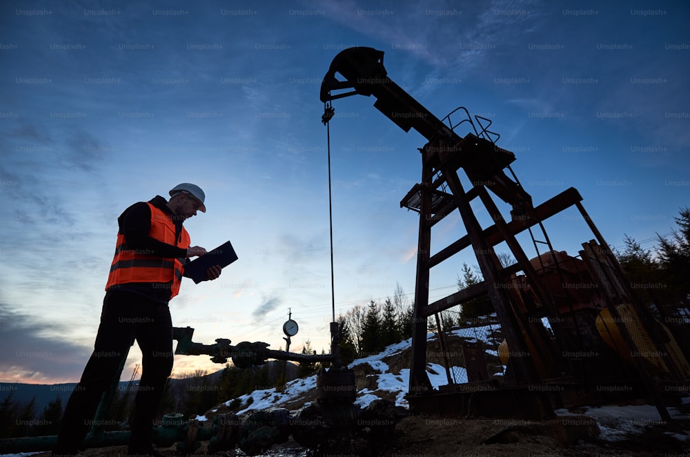 Oil engineer in work vest at oil pump rocker-machine, making notes while checking work of balanced beam petroleum pump jack under beautiful evening sky. Concept of oil extraction, petroleum industry.