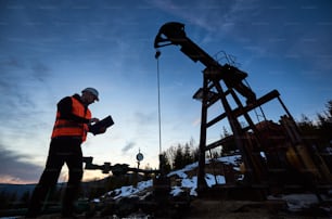 Ingénieur pétrolier en gilet de travail à la culbuteuse de pompe à huile, prenant des notes tout en vérifiant le travail du vérin de pompe à pétrole à faisceau équilibré sous un beau ciel du soir. Concept d’extraction pétrolière, industrie pétrolière.