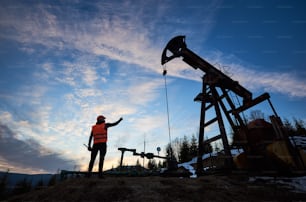 Back view of oil man in work vest standing near petroleum pump jack with sunset on background. Oil worker pointing finger at oil pump rocker-machine. Concept of oil extraction and petroleum industry.