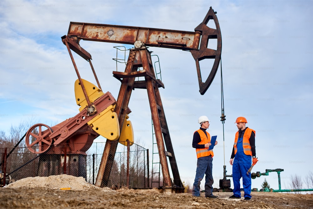 Zwei Erdölingenieure mit Helmen und Arbeitswesten stehen auf dem Territorium eines Ölfeldes mit Ölbohrlochpumpenheber und Himmel im Hintergrund. Ölmann mit Klemmbrett und Gespräch mit Kollege auf dem Ölfeld.