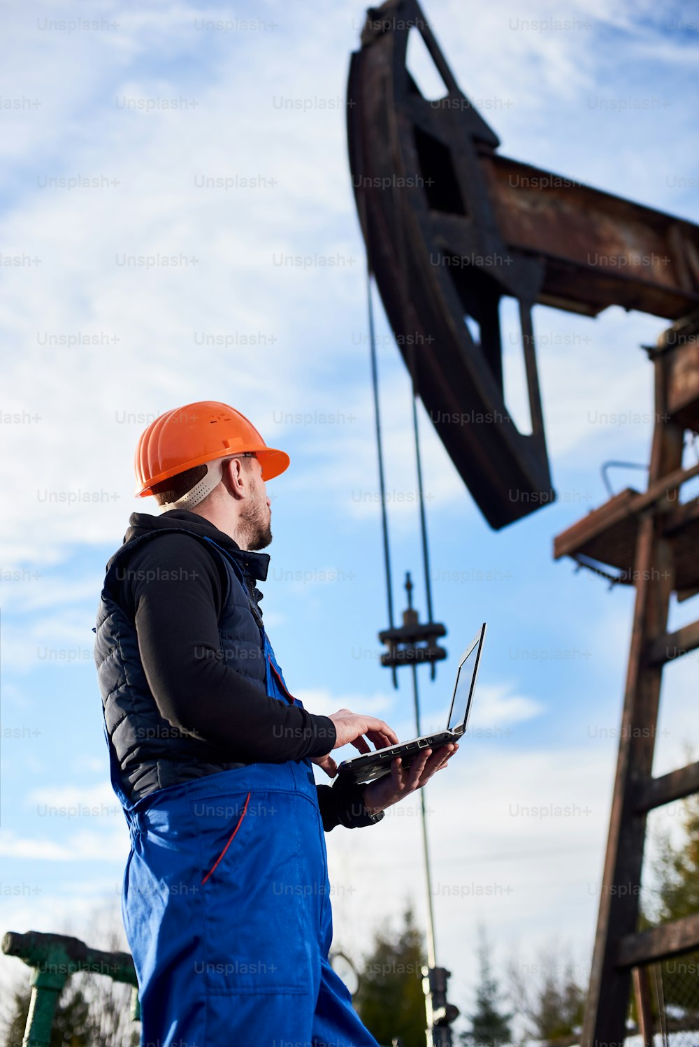 Oil man holding notebook and looking at oil pump rocker-machine. Male worker in uniform and helmet using laptop and controlling work of pump jack. Concept of petroleum industry and oil extraction.
