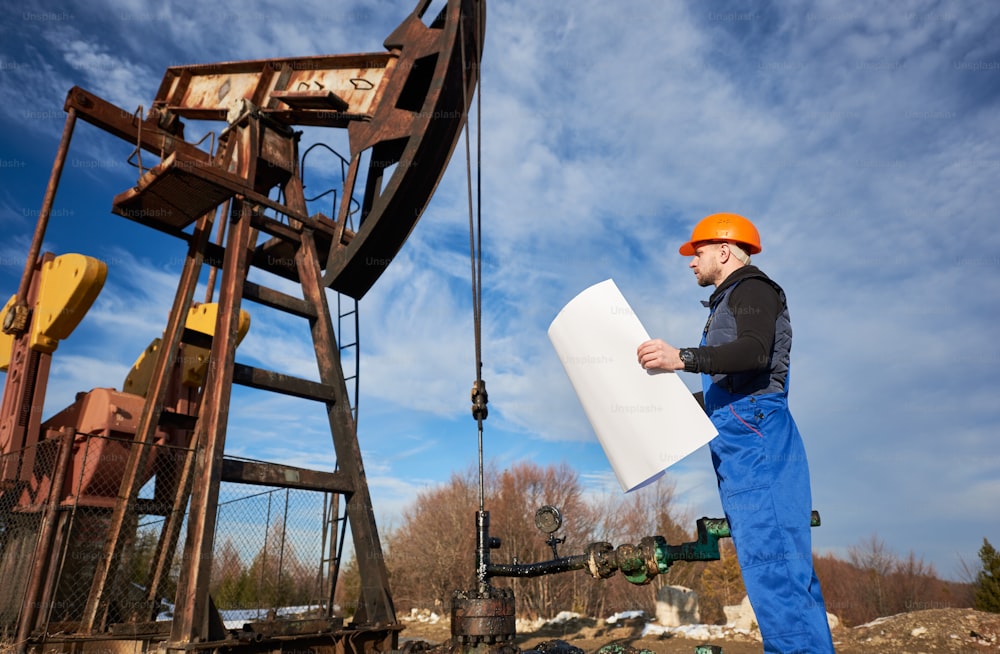 Seitenansicht eines Ölarbeiters, der einen Plan des Ölfeldes an der Ölpumpe hält. Bohrer in Arbeitsuniform und Helm steht in der Nähe der Ölpumpe. Konzept der Erdölindustrie und der Erdölförderung.