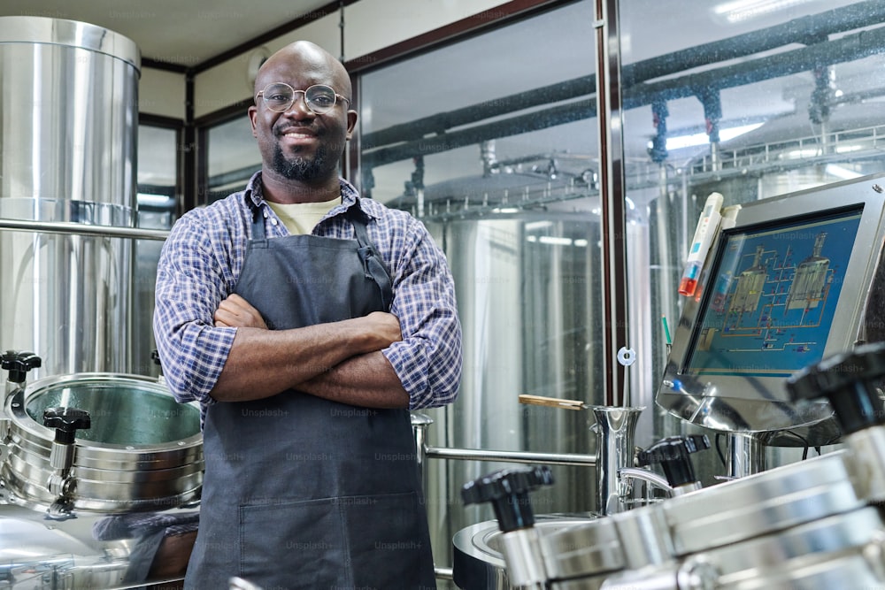 Retrato del ingeniero afroamericano sonriendo a la cámara de pie con los brazos cruzados, trabajando en una cervecería moderna